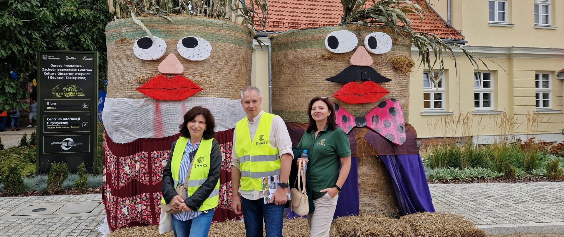 Trzech inspektorów IJHARS stoi na Dożynkach Wojewódzkich; w tle widoczne są tradycyjne ozdoby dożynkowe wykonane ze słomy.