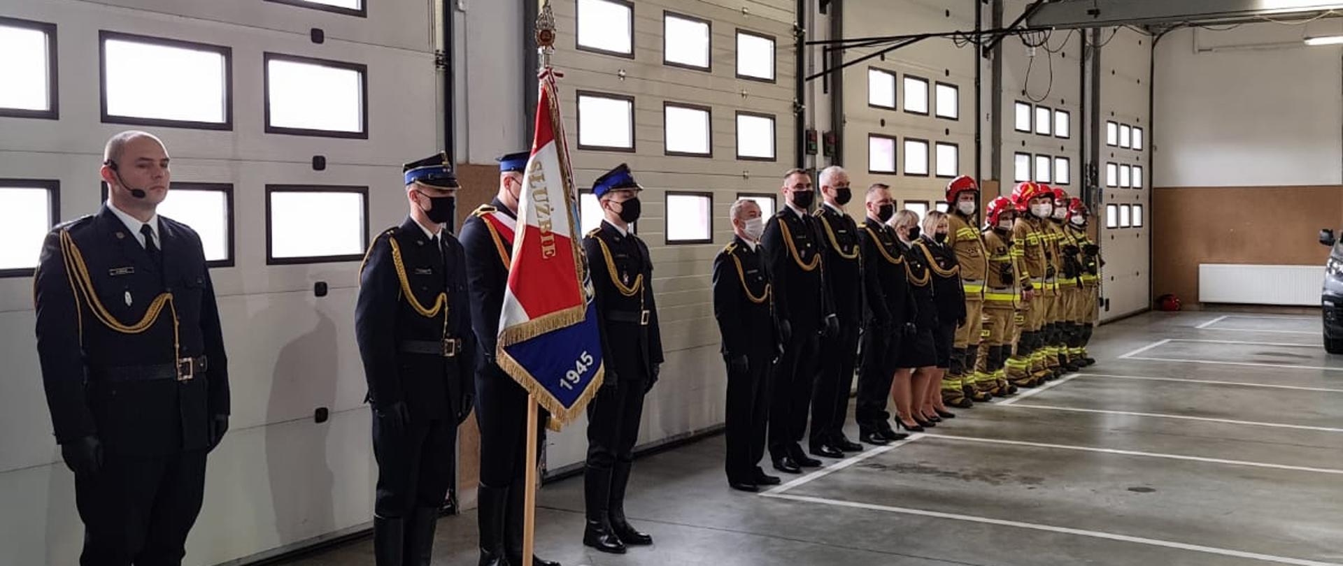 Fotografia przedstawia strażaków stojących na zbiórce w garażu Komendy Powiatowej Państwowej Straży Pożarnej w Malborku.