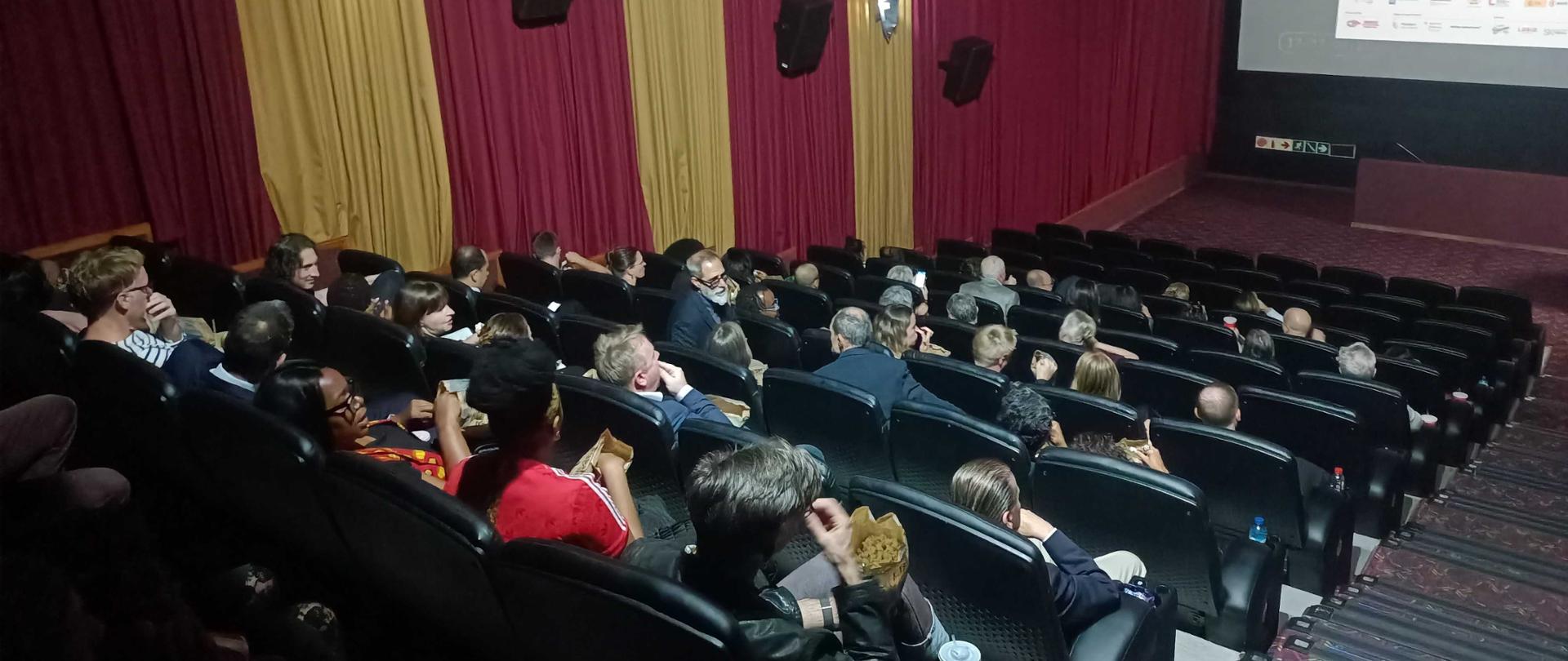 Interior of a cinema auditorium; photo taken at the entrance to the auditorium; a large group of audience members is seated in the left and center sections, eating popcorn; stairs are visible on the right side of the photo; in the background, a screen displays the poster for the European Film Festival; the lights are on; on the left, there are striped drapes in burgundy and gold, and speakers and lights are mounted on the wall;