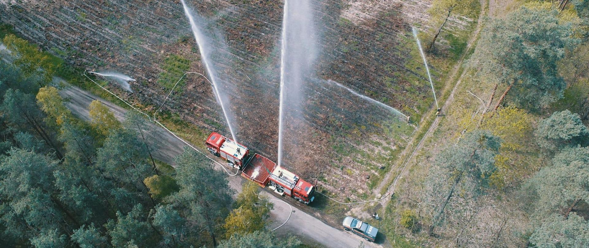 Widok z góry dwóch pojazdów gaśniczych na terenie leśnym stojących przy rozkładanym zbiorniku z wodą, Z pojazdów gaśniczych podawane są prądy gaśnicze z działek. Ponadto podawane są 3 prądy gaśnicze przez strażaków.
