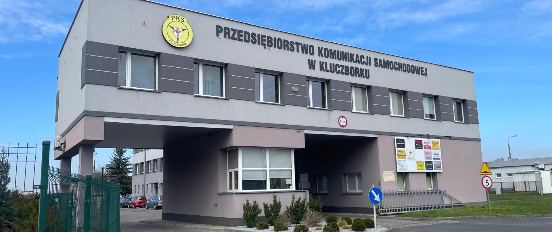  Zdjęcie przedstawia nowoczesny, dwupiętrowy budynek o szarej i jasnofioletowej elewacji. W górnej części fasady znajduje się logo PKS oraz napis „Przedsiębiorstwo Komunikacji Samochodowej w Kluczborku”. W centralnej części budynku znajduje się otwarty przejazd. Po prawej stronie widoczne są tablice informacyjne firm. Przed budynkiem stoją znaki drogowe, w tym ograniczenie prędkości do 5 km/h. Wokół znajduje się ogrodzenie, krzewy i utwardzona droga.