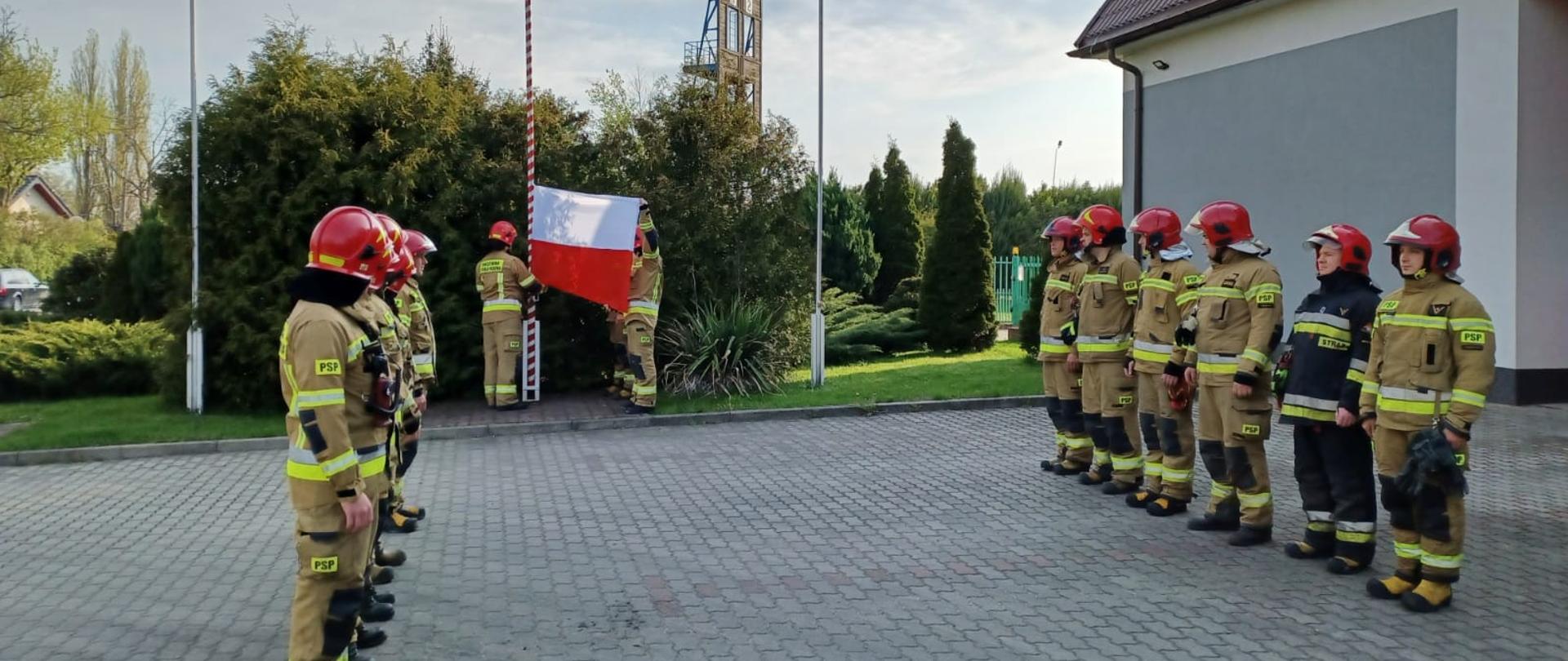Na zdjęciu widać dwie grupy strażaków podczas uroczystej zmiany służby. Wszyscy strażacy maja założone czerwone hełmy. Jeden strażak ubrany jest w ciemny strój a reszta w żółte. Strażacy stoją w dwóch rzędach naprzeciwko siebie. W tle widać strażaków w żółtych strojach i czerwonych hełmach, którzy zawieszają flagę Polski na maszcie. Uroczysta zbiórka odbywa się na placu manewrowym przed budynkiem Komendy Powiatowej Państwowej Straży Pożarnej w Łęczycy. W tle widać krzewy. Jest widno.