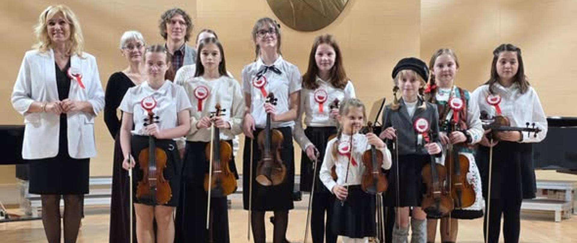 Zdjęcie przedstawia grupę uczniów stojącą z nauczycielami na scenie sali koncertowej. Uczniowie w dłoniach trzymają skrzypce. 