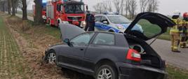 Na zdjęciu widać rozbity pojazd osobowy oraz na drugim planie wóz strażacki i strażaków 