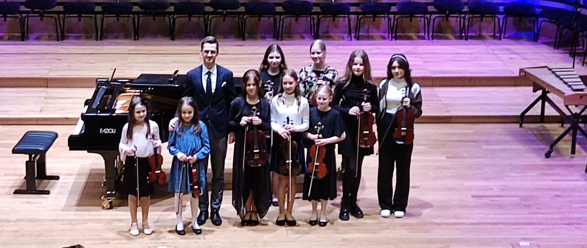 Dziewiątka dziewczyn stoi w dwóch rzędach razem z nauczycielem, za nimi stoi fortepian.