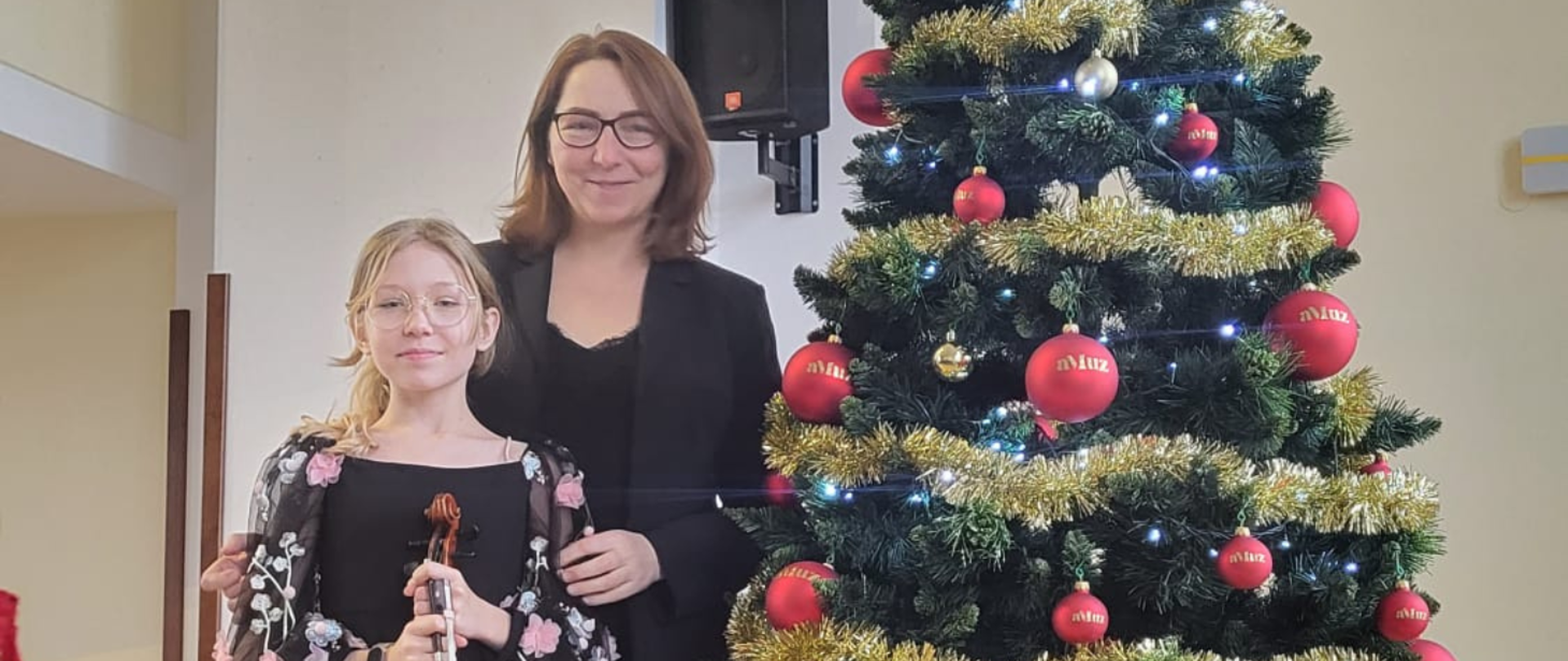 Na zdjęciu uczennica blondynka w okularach w czarnej sukience, która trzyma skrzypce. W tle za nią kobieta w okularach, brązowych włosach, w czarnej marynarce.Obok nich stoi choinka, a na niej czerwone bombki ze złotym napisem "aMuz" i złote łańcuchy.