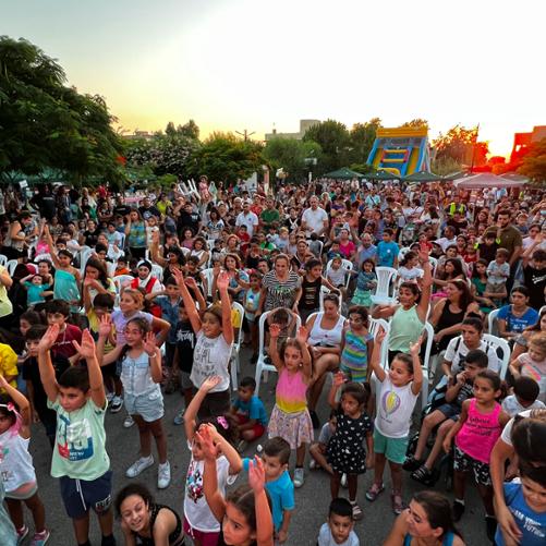 Duża grupa dzieci zebrana na placu wyciąga ręce do góry, z tyłu za dziećmi stoją dorośli 