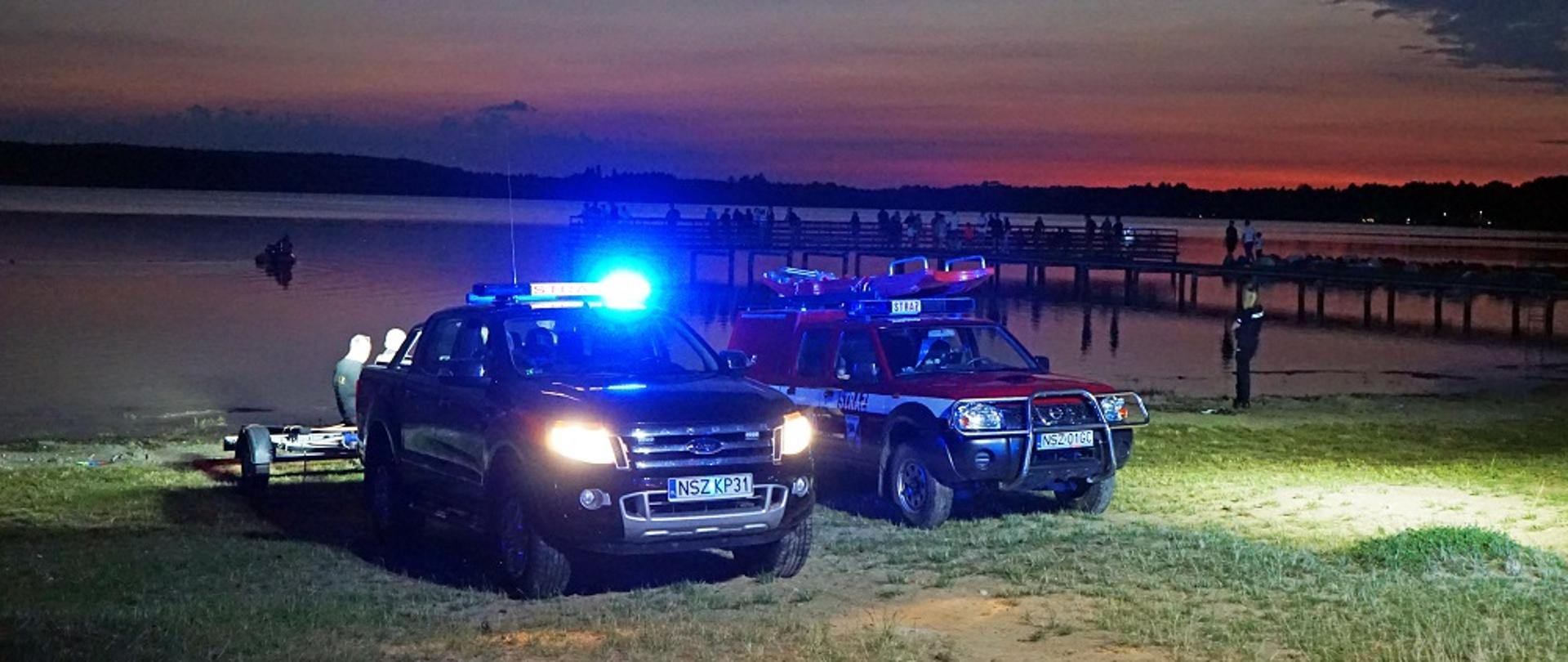 Zdjęcie przedstawia strażaków oraz samochody ratowniczo-gaśnicze prowadzące poszukiwania zaginionych osób na jeziorze Świętajno.