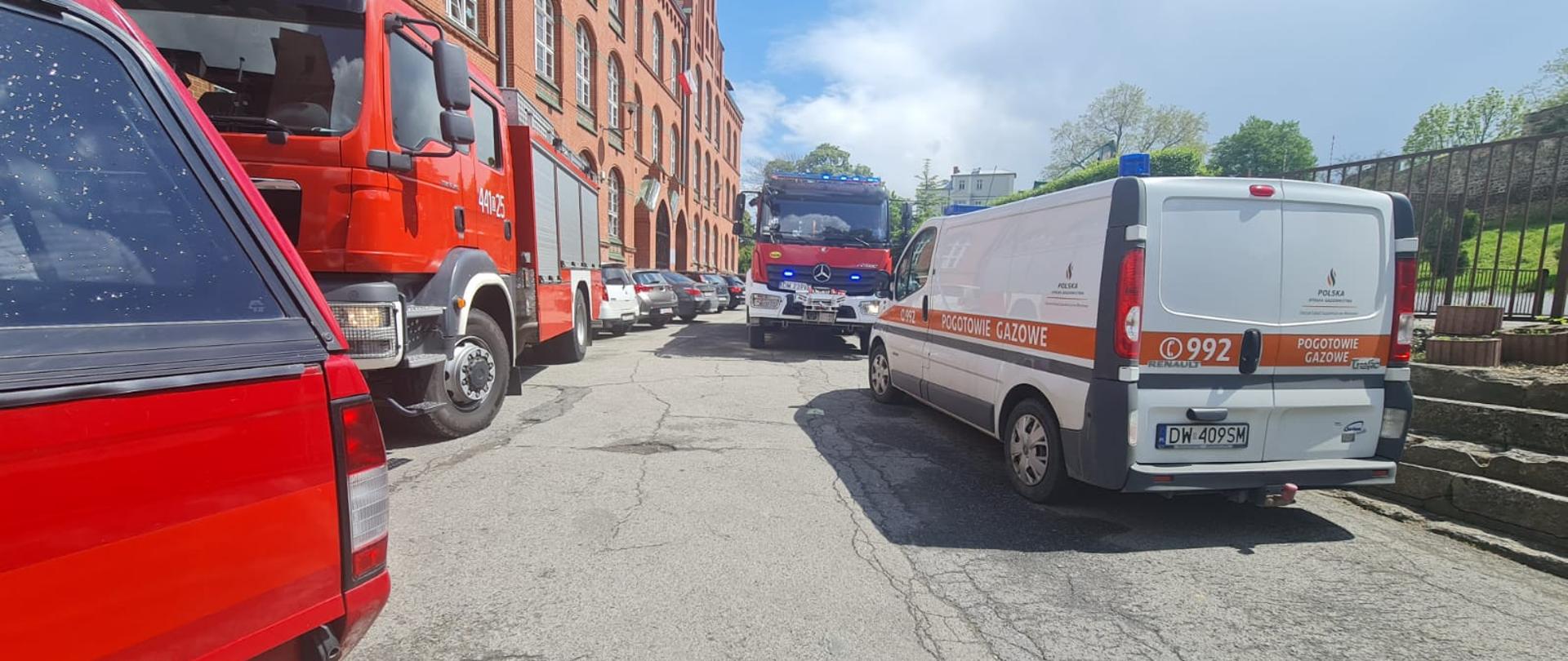 Obraz przedstawia pojazdy pożarnicze oraz pojazd pogotowia gazowego. W tle samochody osobowe i budynek szkoły.