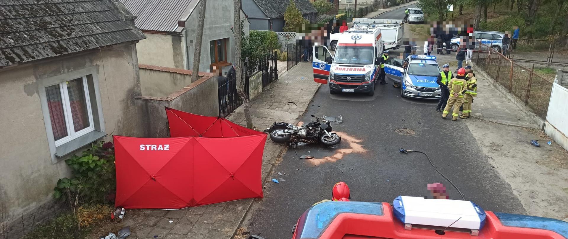 Na ulicy lezy motocykl przed nim widoczny przód samochodu strażackiego przy budynku po lewej stronie rozstawiony czerwony parawan , za motocyklem stoi karetka pogotowia oraz radiowóz policji przy radiowozie dwóch strażaków i policjant za karetką i radiowozem osoby postronne.