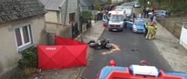 Na ulicy lezy motocykl przed nim widoczny przód samochodu strażackiego przy budynku po lewej stronie rozstawiony czerwony parawan , za motocyklem stoi karetka pogotowia oraz radiowóz policji przy radiowozie dwóch strażaków i policjant za karetką i radiowozem osoby postronne.