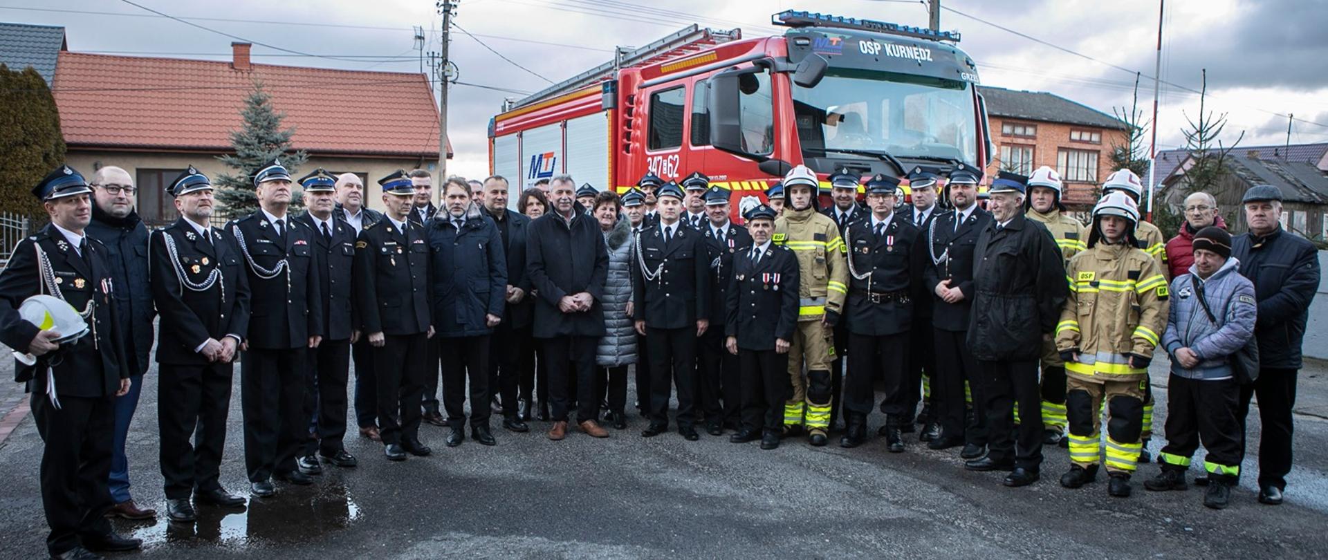 Strażacy OSP i PSP oraz zaproszeni goście pozują do zdjęcia na tle samochodu gaśniczego oraz domów