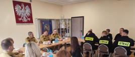 Na fotografii widoczny Opolski Komendant Wojewódzki Państwowej Straży Pożarnej st. bryg. Paweł Kielar zasiadający wspólnie przy stole z kierownictwem, funkcjonariuszami i pracownikami KP PSP w Oleśnie na sali konferencyjnej. W tle widoczny wizerunek Godła RP.