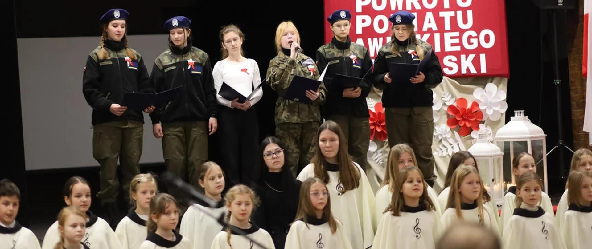  Na scenie w ośrodku kultury w Jutrosinie występują uczennice klasy mundurowej. W rękach trzymają teczki z tekstem pieśni Jedna z nich, bez nakrycia głowy śpiewa, trzymając w prawej ręce mikrofon. Z prawej strony na scenie znajduje sie dekoracja - na czerwonym tle widnieje biały napis: 104. Rocznica Powrotu Powiatu Rawickiego do Polski. Przed sceną w kremowych pelerynach z kluczem wiolinowym na przodzie, w dwóch rzędach stoją dziewczęta tworzące chórek.