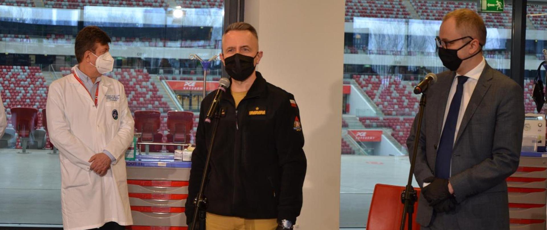 Na zdjęciu 3 osoby w maseczkach: jedna w garniturze, jedna w mundurze strażackim, jedna w białym fartuchu. Przed dwiema z nich mikrofony na stojakach. W tle za przeszkleniem siedzenia na trybunach stadionu.