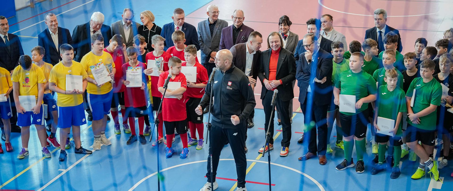 Konferencja ministra sportu i turystyki Jakuba Rutnickiego na sali gimnastycznej. Minister mówi do mikrofonu. Za nim stoją pozostali uczestnicy spotkania: wojewoda mazowiecki Mariusz Frankowski, wicemarszałek województwa mazowieckiego Wiesław Raboszuk, wójt gminy Załuski Kamil Koprowski, poseł Adam Krzemiński, przedstawiciele samorządów oraz dzieci w strojach sportowych. Z lewej strony, nieco w tyle widać drabinki gimnastyczne. Zdjęcie robione jest z góry, częściowo przez siatkę.