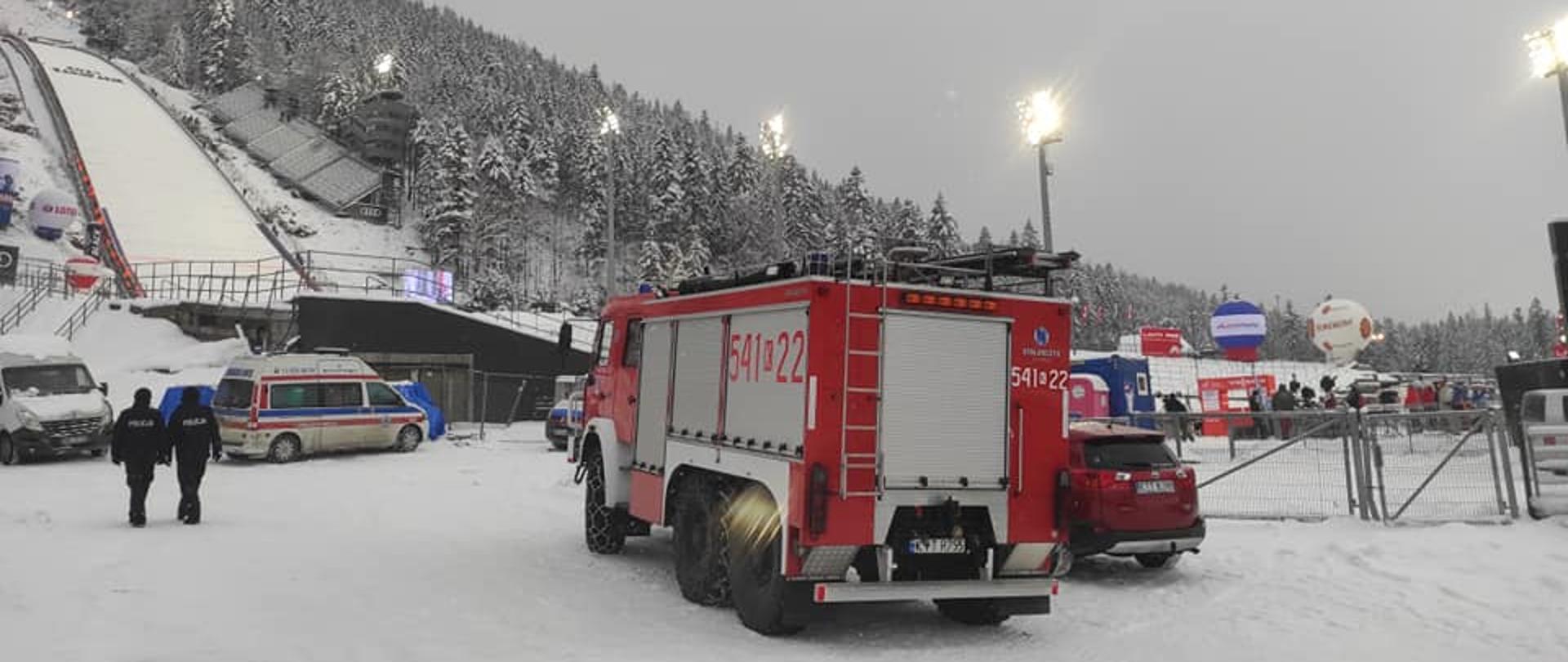 W centralnej części pojazd straży pożarnej na tle ośnieżonej skoczni narciarskiej. Po lewej stronie dwóch policjantów.