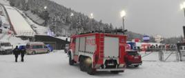 W centralnej części pojazd straży pożarnej na tle ośnieżonej skoczni narciarskiej. Po lewej stronie dwóch policjantów.