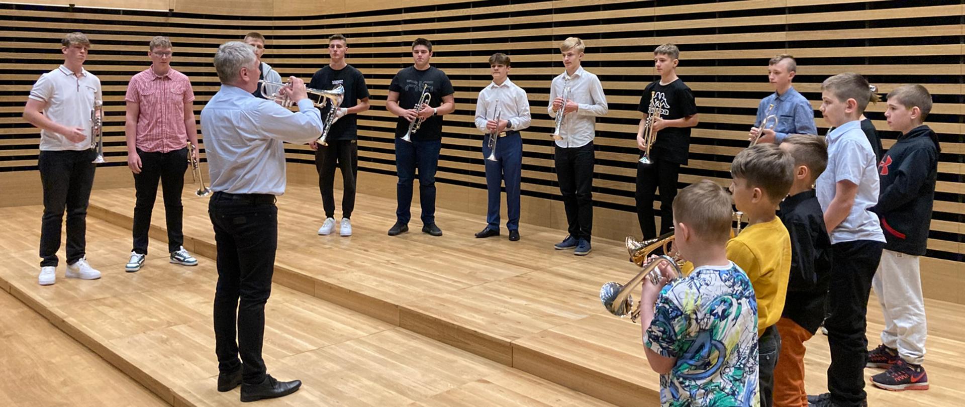 Mężczyzna stoi i gra na trąbce, wokół niego w półkolu stoi czternastu trębaczy (dzieci i młodzież) i trzymają trąbki przed sobą.