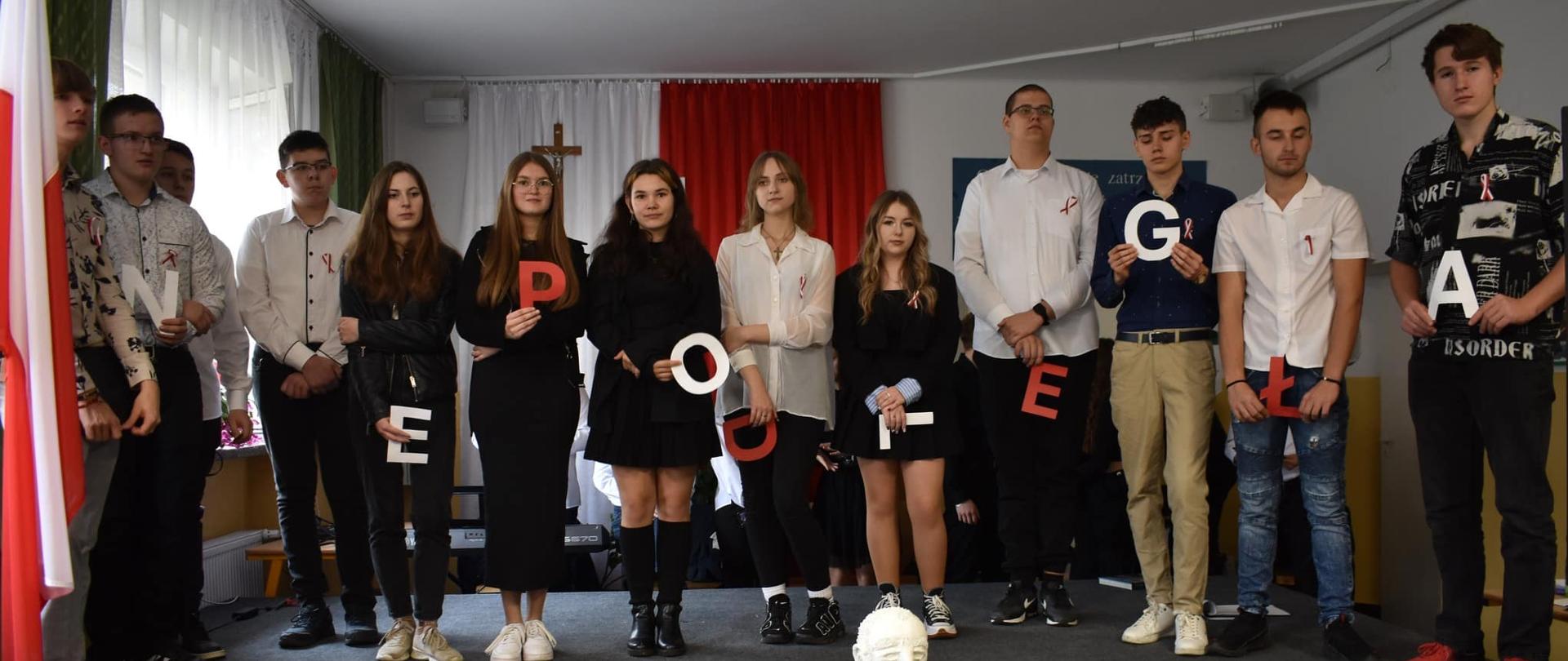 Grupa uczniów na scenie podczas uroczystości. Trzymają duże litery, które tworzą napis "NIEPODLEGŁA". Na środku sceny stoi popiersie Józefa Piłsudskiego, a w tle wisi biało-czerwona flaga.