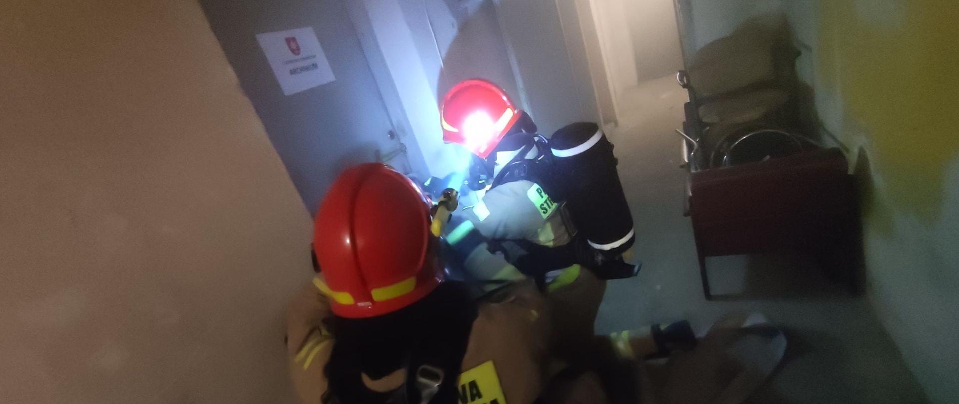 NA ZDJĘCIU DWÓCH STRAŻAKÓW, STRAŻAK BĘDĄCY NA PIERWSZYM PLANIE JEST ODWRÓCONY PLECAMI, STRAŻACY MAJĄ NA GŁOWACH CZERWONE KASKI I BUTLE Z TLENEM NA PLECACH. WSZĘDZIE UNOSI SIĘ SIWY DYM, JEST SŁABA WIDOCZNOŚĆ. STRAŻACY PRÓBUJĄ OTWORZYĆ DRZWI , NA KÓRYCH WIDNIEJE NAPIS ARCHIWUM.
