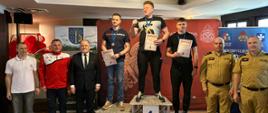 Kolorowa fotografia wykonana w pomieszczeniu. Przedstawia strażaków - pływaków w strojach sportowych na podium podczas wręczania dyplomów i medali po mistrzostwach województwa w pływaniu..Z boku podium komendanci wręczający nagrody. W tle banery Państwowej Straży Pożarnej.