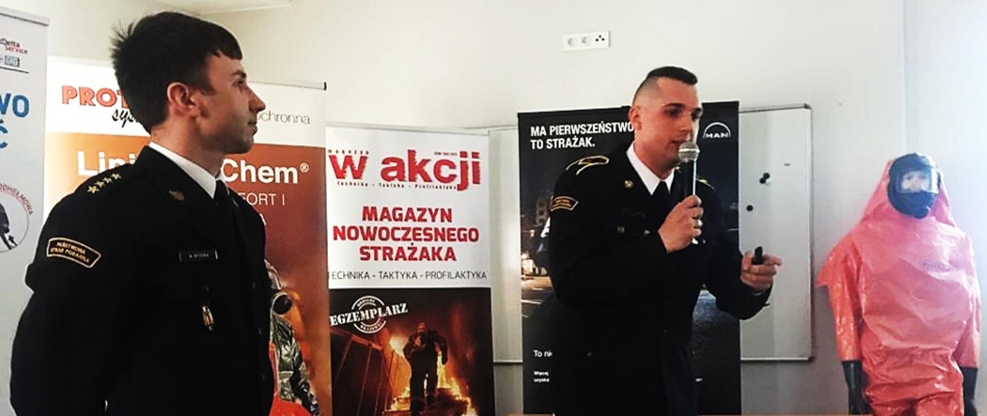 Na pierwszym planie stojący strażak w mundurze galowym bez sznura, w tle drugi strażak w mundurze galowym bez sznura trzymający mikrofon. Za nimi banery reklamowe. W głębi manekin w kombinezonie ochronnym