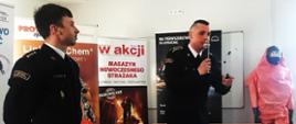 Na pierwszym planie stojący strażak w mundurze galowym bez sznura, w tle drugi strażak w mundurze galowym bez sznura trzymający mikrofon. Za nimi banery reklamowe. W głębi manekin w kombinezonie ochronnym