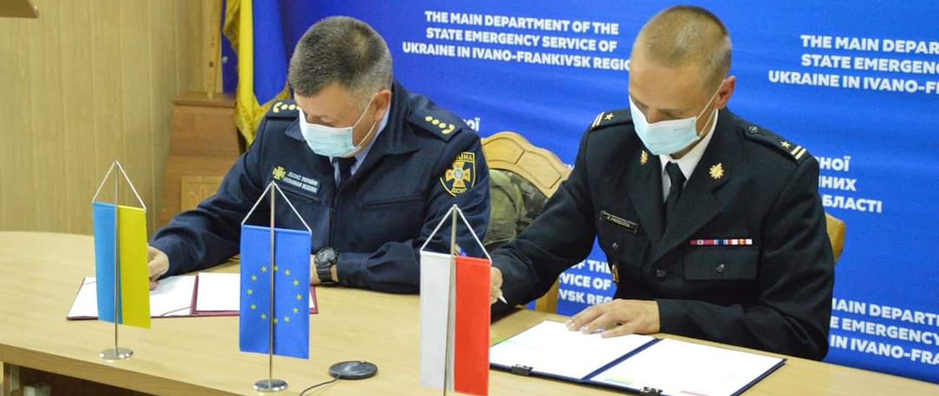 przy stole siedzą 2 osoby, które podpisują dokumenty. Na stole znajdują się flagi Polski i Ukrainy;