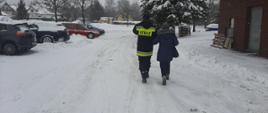 Na zdjęciu widoczny strażak na zaśnieżonej drodze prowadzący kobietę pod rękę. W oddali widoczny na parkingu samochód strażacki koloru czerwonego którym kobieta zostanie przetransportowana do punktu szczepień.