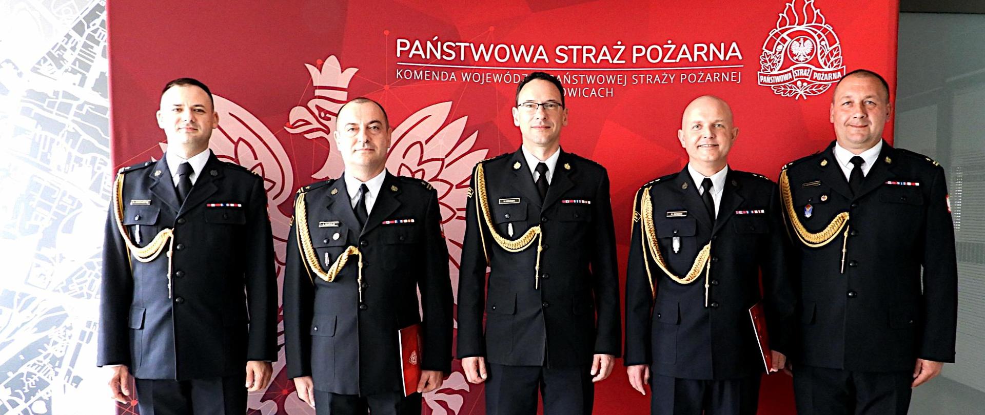Na tle scianki koloru czerwonego z orłem w koronie i napisem u góry Państwowa Straż Pożarna Komenda Wojewódzka Państwowej Straży Pożarnej w Katowicach, stoją w mundurach wyjściowych od prawej komendant miejski PSP w Bytomiu mł. bryg. Tomasz Rytkowski, zastęcpca komendanta kpt. Rafał Borzek, w środku śląski komendant wojewódzki PSP st. bryg. Wojciech Kruczek, zastępca komendanta miejskiego PSP w Dąbrowie Górniczej mł. bryg. Łukasz Musialik i komendant miejski w Dąbrowie Górniczej 