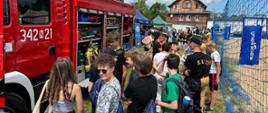 Młodzież szkolna z plecakami zgromadziła się do około pojazdu pożarniczego. Wśród nich znajduje się strażak, który prezentuje pojazd strażacki.