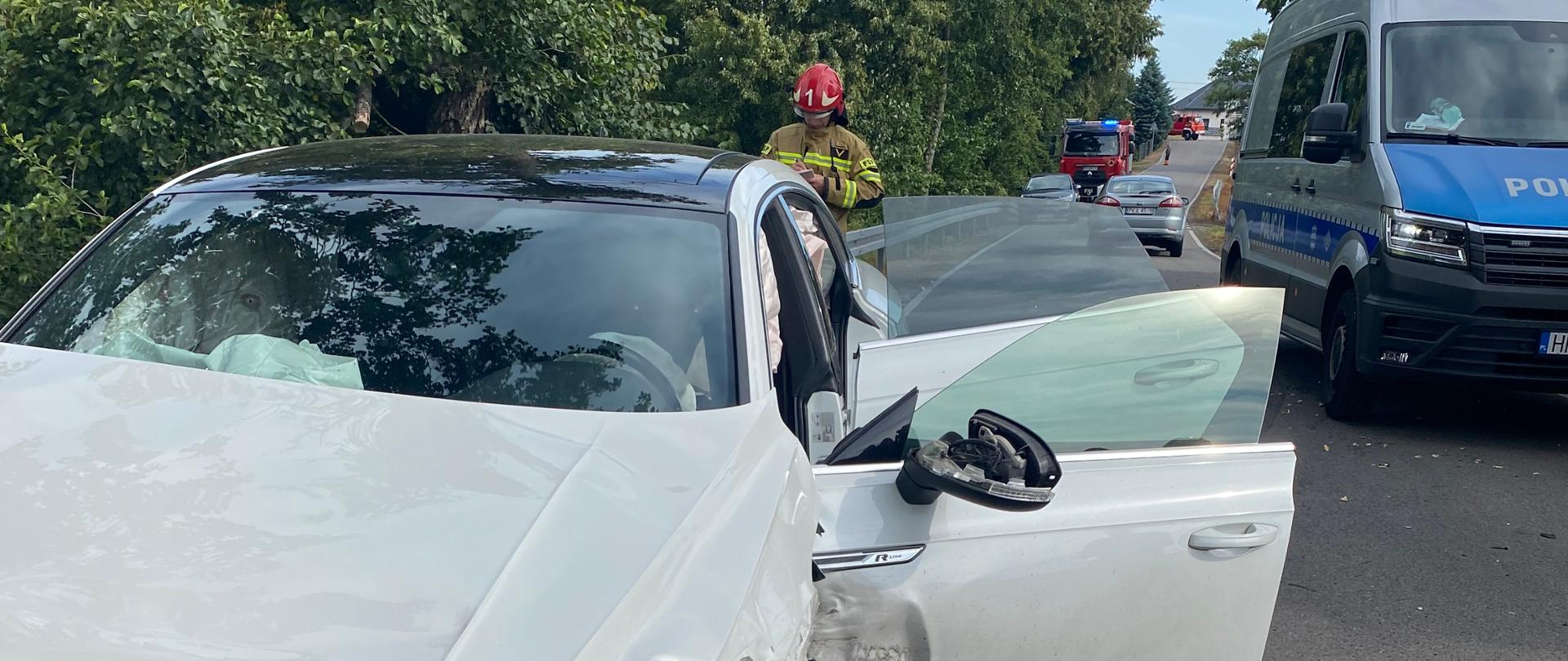 Na zdjęciu widać uszkodzony pojazd na drodze publicznej. Droga asfaltowa. Zdjęcie wykonane w ciągu dnia. Widać zarośla w kolorze zielonym oraz bariery ochronne drogi. Widać pojazdy służb mundurowych. Posiadają sygnalizację ostrzegawczą. Na miejscu funkcjonariusze.