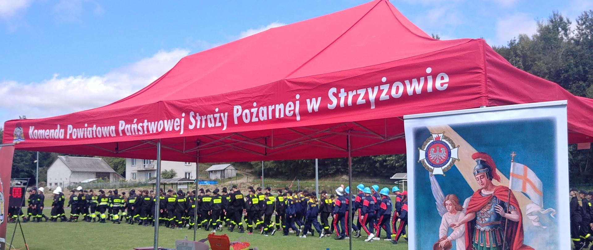 Na pierwszym planie widać czerwony namiot pod którym stoją stolik. Po prawej stronie widać baner ze św. Florianem a po lewej baner KP PSP w Strzyżowie. W tle widać kolumnę uczestników wchodzących na stadion