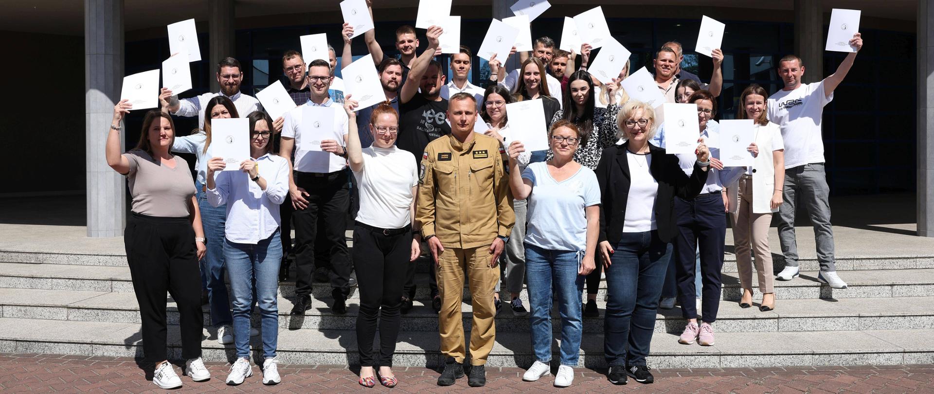 Grupa uczestników szkolenia inspektorów ochrony przeciwpożarowej pozuje przed budynkiem Centralnej Szkoły PSP w Częstochowie. Uczestnicy trzymają w rękach zaświadczenia ukończenia szkolenia. W centrum zdjęcia stoi Zastępca Komendanta Centralnej Szkoły PSP w umundurowaniu służbowym