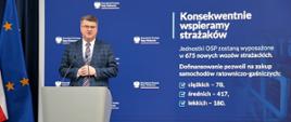 minister Wąsik przemawia z trybuny w tle duży baner z kancelarii prezesa rady ministrów po lewej stronie flaga unii europejskiej