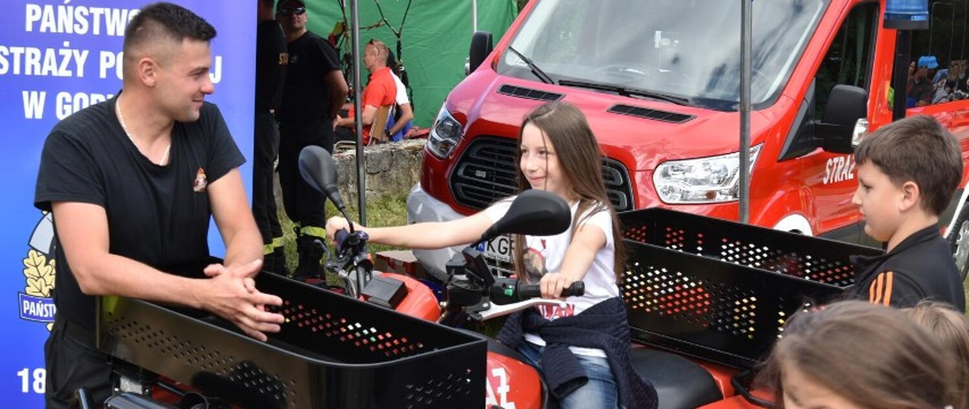 Na zdjęciu widoczna dziewczynka siedząca na quadzie i kilka dzieci uczestniczących w pikniku rodzinnym przed namiotem koloru niebieskiego i banerem z napisem Komenda Powiatowa PSP w Gorlicach.