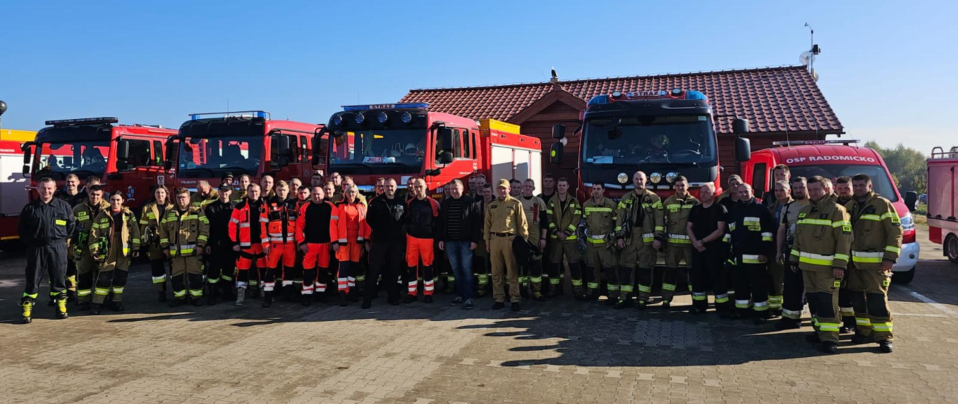 Strażacy i ratownicy medyczni stoją w szeregu na tle pojazdów ratowniczych.