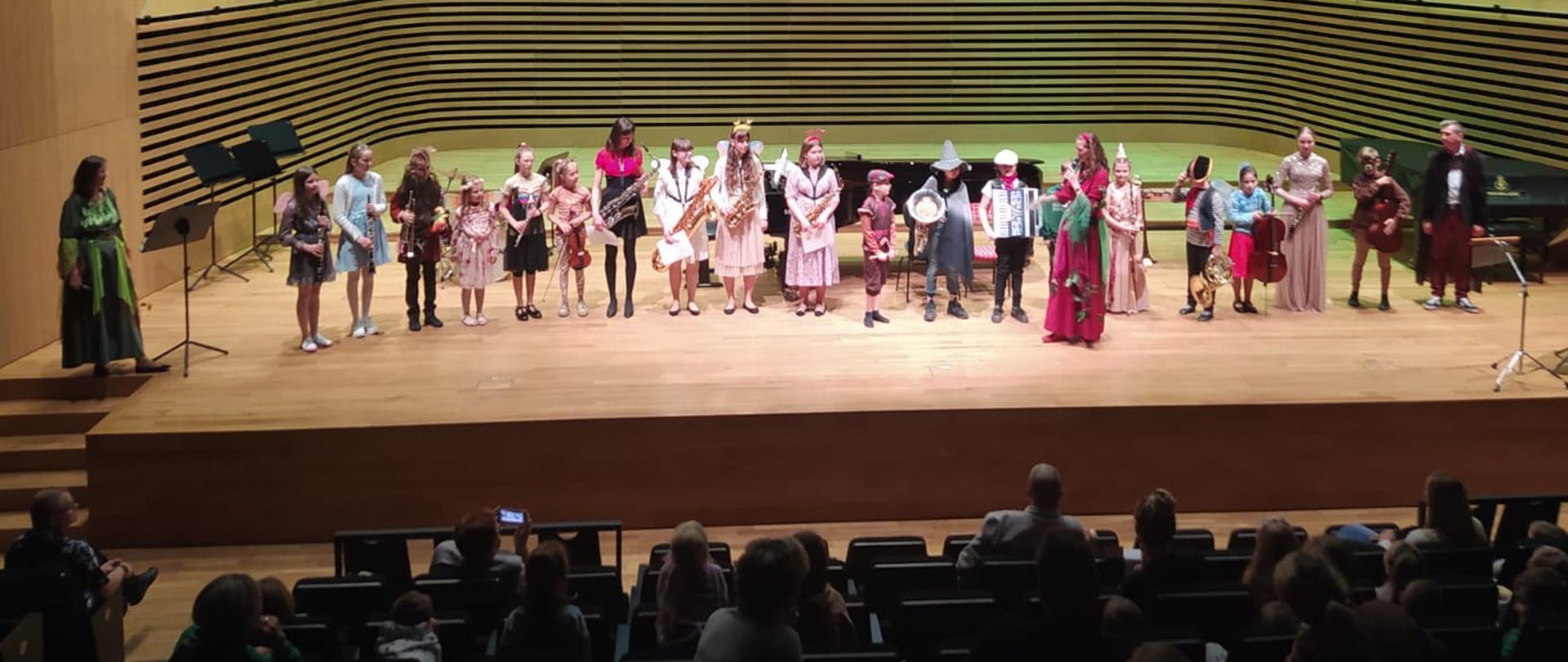 18 dzieci w różnych przebraniach stoi w rzędzie na estradzie sali koncertowej PSM w Jastrzębiu-Zdroju, po lewej stronie stoi dwie kobieta w przebraniu wróżki, po prawej stoi mężczyzna w przebraniu czarodzieja, przed dziećmi stoi kobieta mówiąca do mikrofonu w przebraniu wróżki. Nad estradą widoczny rzutnik z napisem "Zaczarowana muzyczna kraina" Dziękujemy!