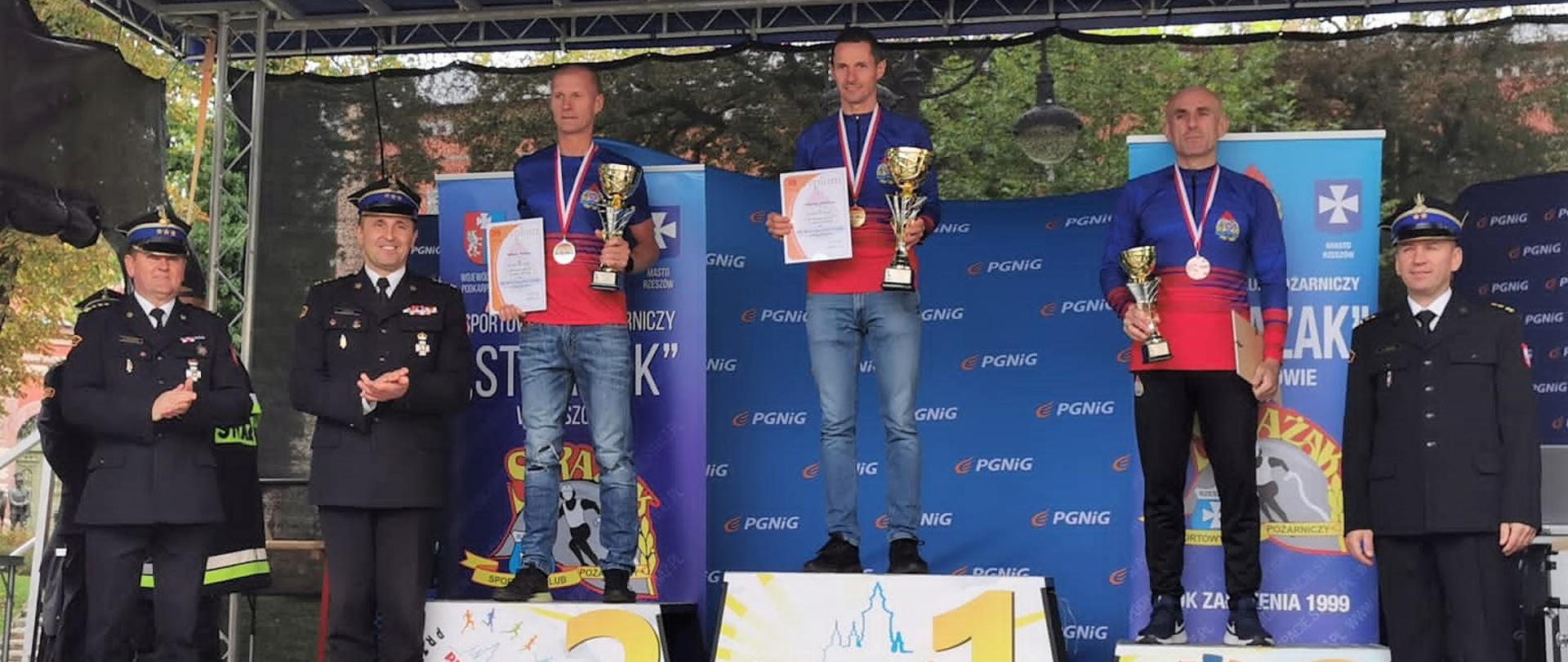 Zdjęcie przedstawia podium, na którym stoją zwycięzcy kategorii C tegorocznych Mistrzostw Polski Strażaków w Biegu Ulicznym.