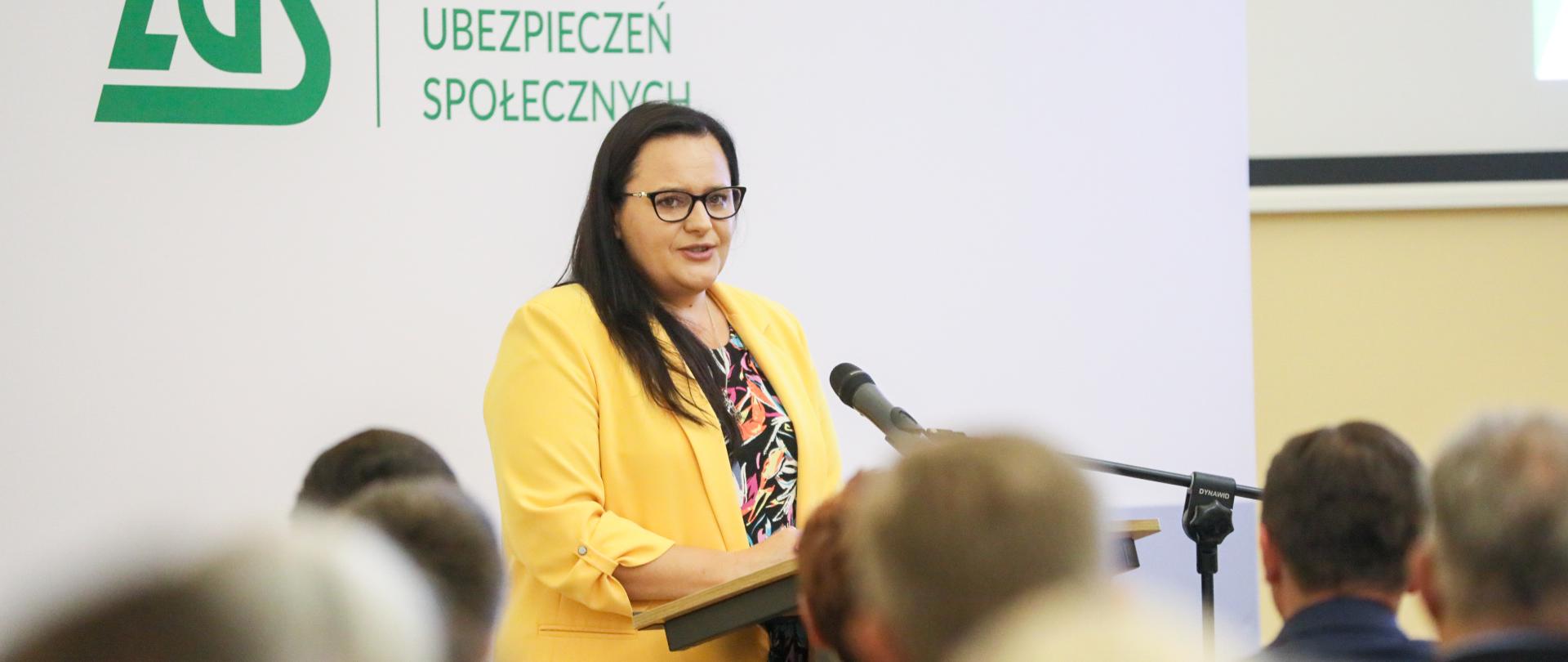 Wiceminister Małgorzata Jarosińska-Jedynak w mównicy na tle ścianki ZUS. Przed nią siedzi publiczność.