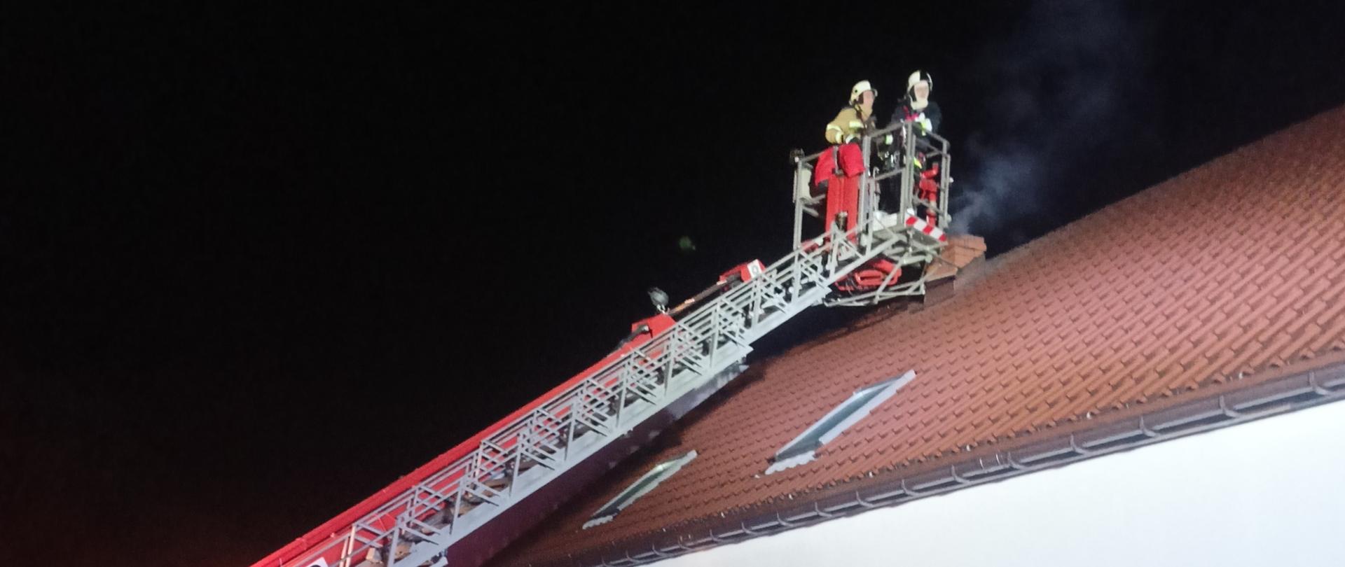 Przy budynku ustawiony podnośnik z wysuniętym ramieniem. W koszu strażacy gaszący pożar sadzy w kominie. Z komina unosi się dym. Pora nocna. 