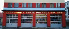 Zdjęcie przedstawia część ściany frontowej budynku Komendy Powiatowej Państwowej Straży Pożarnej w Brzozowie malowaną pasami koloru czerwonego, niebieskiego i żółtego, w okresie zimowym. Na ścianie napis o treści Państwowa Straż Pożarna Tel. 998. Na parterze widoczne cztery bramy wyjazdowe, na pozostałych piętrach okna pomieszczeń.