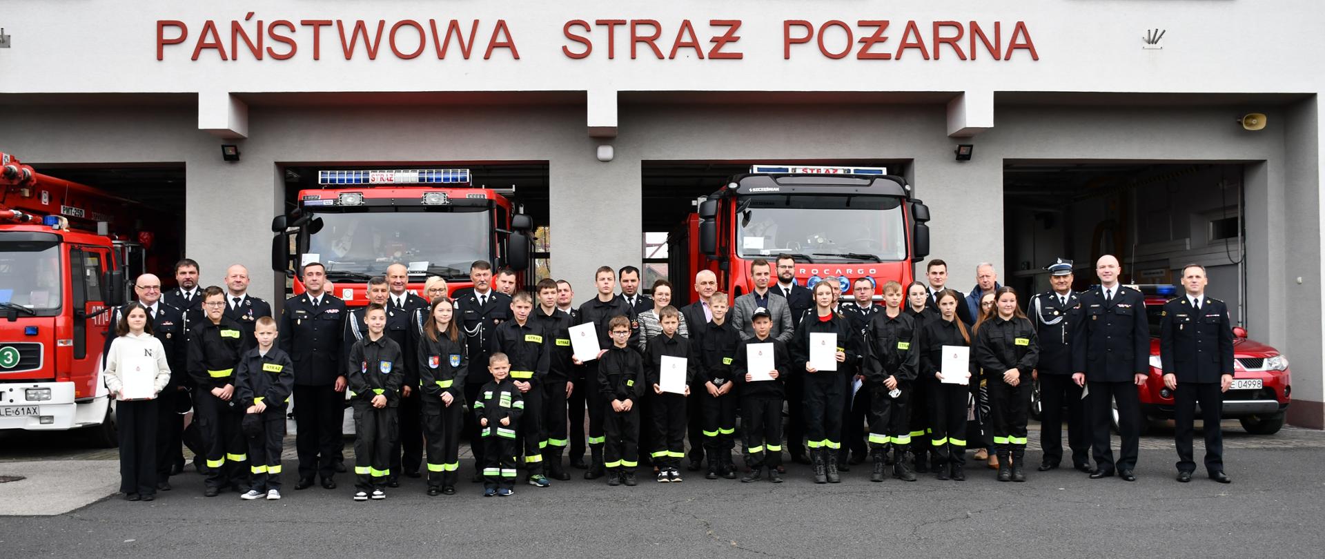 Na zdjęciu widzimy komendantów PSP, władze lokalne, prezesów OSP wraz z dziećmi z młodzieżowych drużyn pożarniczych pozujących do zdjęcia grupowego na tle budynku komendy PSP.