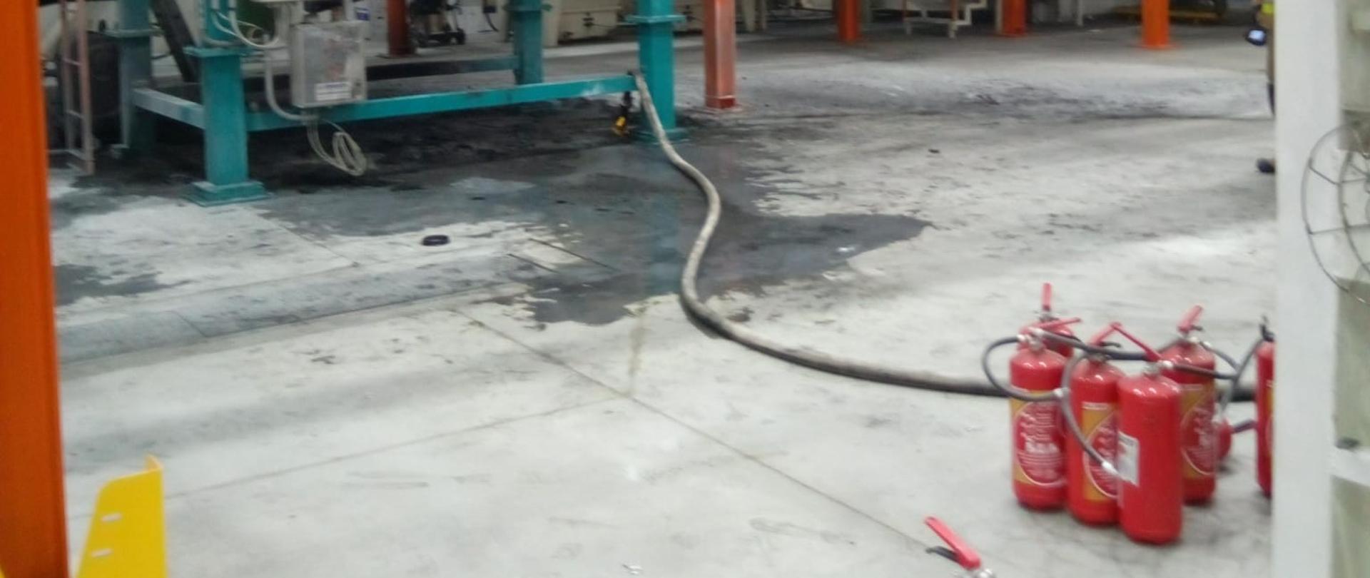 Zdjęcie przedstawia wnętrze hali gdzie doszło do pożaru filtrów wewnętrznych w separatorze odpylającym. Na podłodze leżą i stoją gaśnice proszkowe, którymi pracownicy próbowali ugasić ogień.