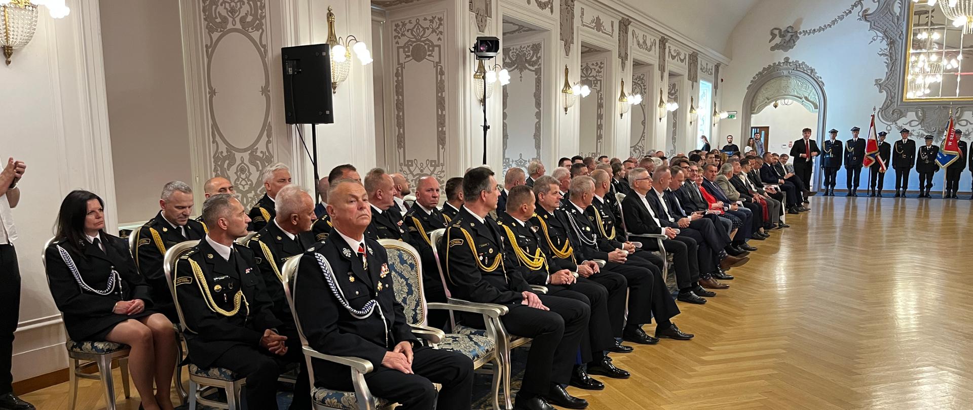 Na zdjęciu widać dużą, elegancką salę podczas uroczystości. Po lewej stronie siedzą strażacy i zaproszeni goście, ustawieni w długim rzędzie. W głębi sali widać poczet sztandarowy oraz osoby prowadzące uroczystość.
