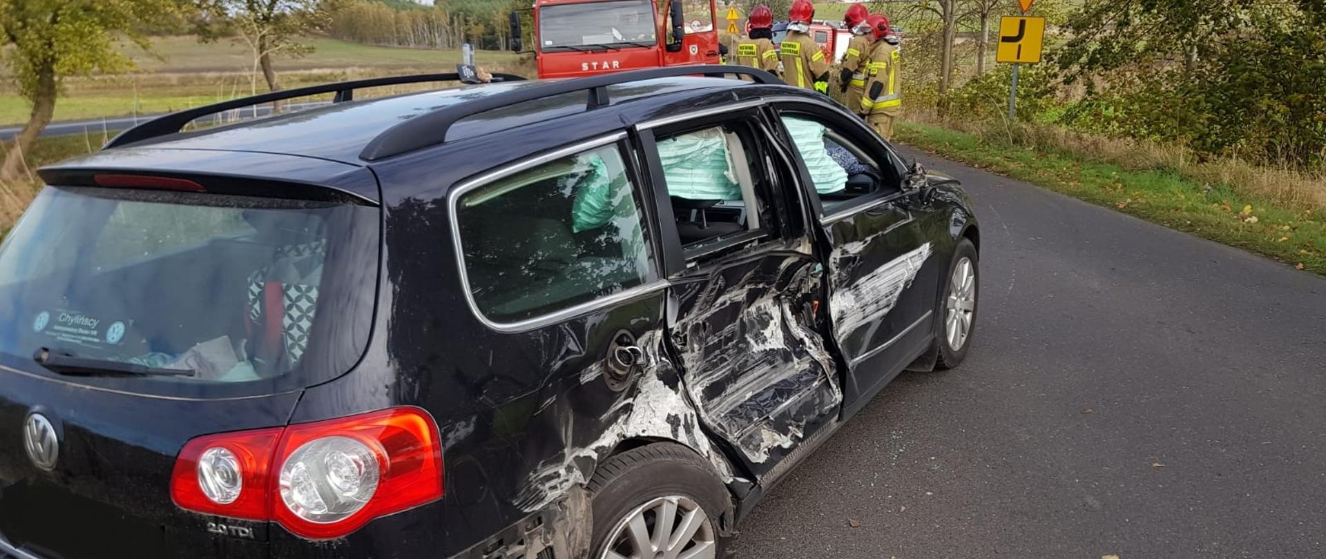 Na zdjęciu widać uszkodzony samochód osobowy biorący udział w zdarzeniu. W pojeździe wystrzeliły poduszki powietrzne i uszkodzony jest cały prawy bok pojazdu. W tle widać samochód strażacki przy którym stoją strażacy