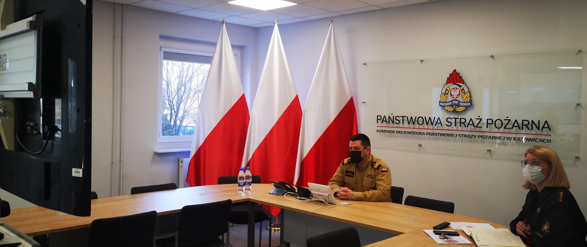 Na zdjęciu sala sztabowa. Śląski Komendant Wojewódzki PSP prowadzi wideokonferencję z kadrą kierowniczą PSP woj. śląskiego. Po prawej stronie siedzi Naczelnik Wydziału Organizacyjnego. W tle po lewej stronie ustawione trzy flagi narodowe. Na ścianie napis Państwowa Straż Pożarna i logo PSP.