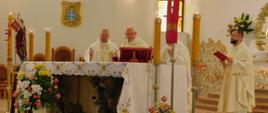 Na zdjęciu widoczni są księża podczas odprawiania mszy św. Księża ubrani są w ornaty koloru białego. Masza odprawiana jest na ołtarzu przykrytym białym obrusem. Na pierwszym planie widoczny jest bukiet kwiatów świece i krzyż.