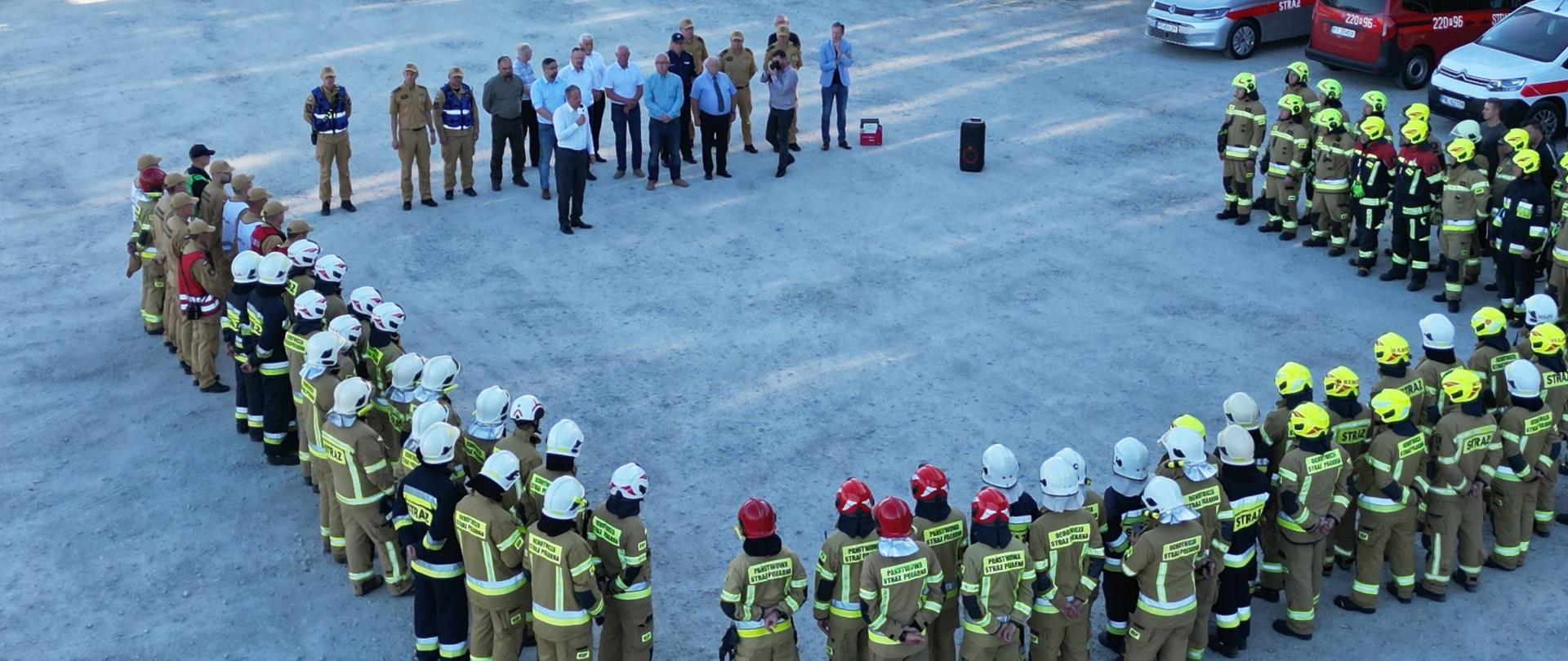 Zbiórka końcowa, strażacy stoją w kształcie litery U, na froncie przemawiają zaproszeni goście